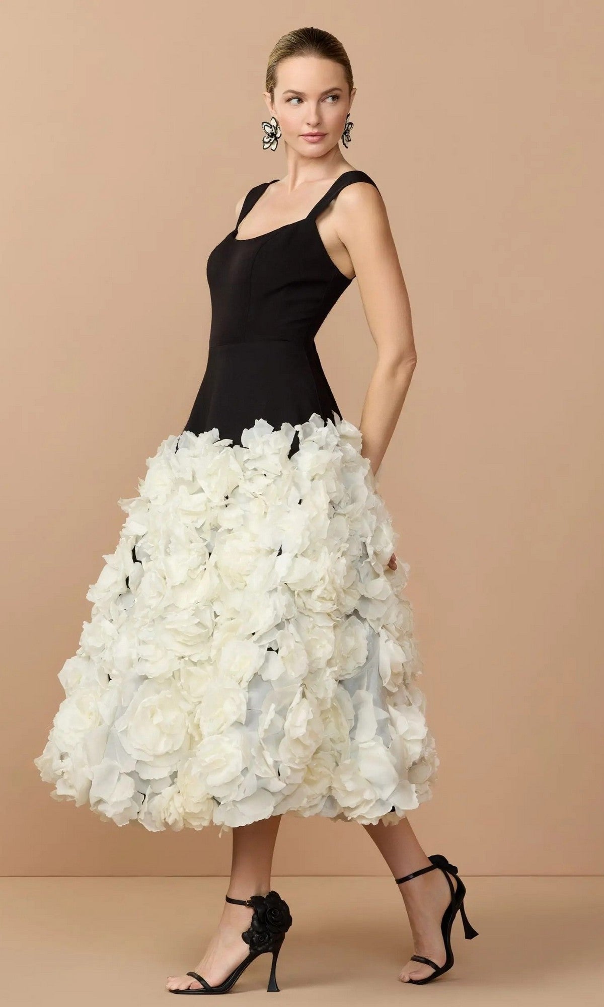A woman stands against a beige background in the Mac Duggal 21080 midi wedding guest dress—featuring a sleeveless black bodice and voluminous white skirt with 3D organza flowers—paired with black heels and floral earrings.